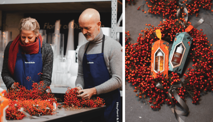 Two people making rosehip Christmas wreath in workshop, with finished wreaths featuring red rosehip berries on dark background