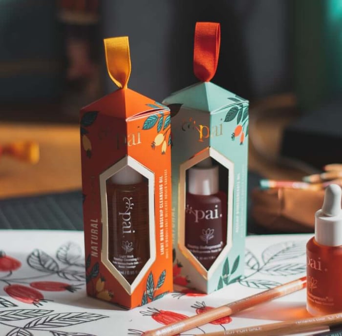 Two Pai Skincare Natural Treasures Christmas gift boxes with botanical illustrations, containing Rosehip BioRegenerate Oil and Light Work Cleansing Oil, displayed with artistic materials