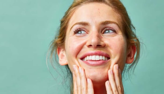 Smiling woman with radiant glowing skin gently touching her face against mint green background