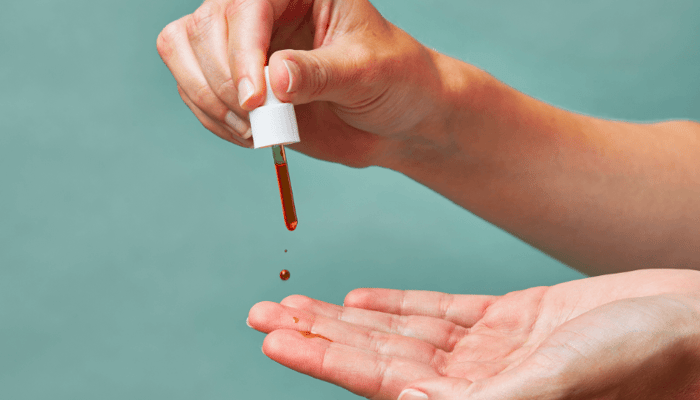 Hand dispensing drops of Pai Rosehip BioRegenerate Oil from dropper bottle onto palm against sage green background