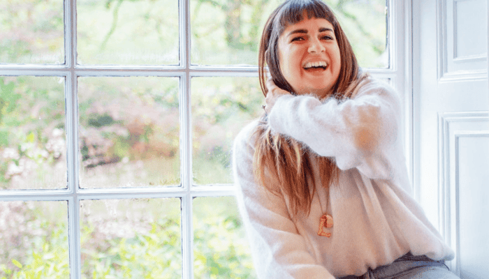 Life Coach and Comparison Coach Lucy Sheridan smiling warmly by a bright window, embodying the radiant glow of self-care and wellbeing