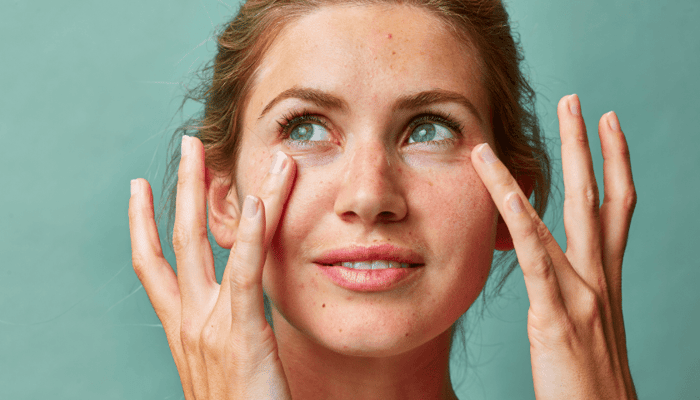 Young woman gently touching temples with fingertips, demonstrating face massage technique on mint green background