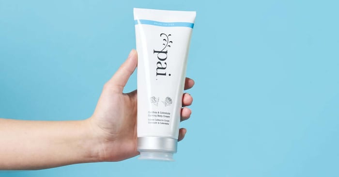 Hand holding Pai Comfrey & Calendula Calming Body Cream tube against light blue background