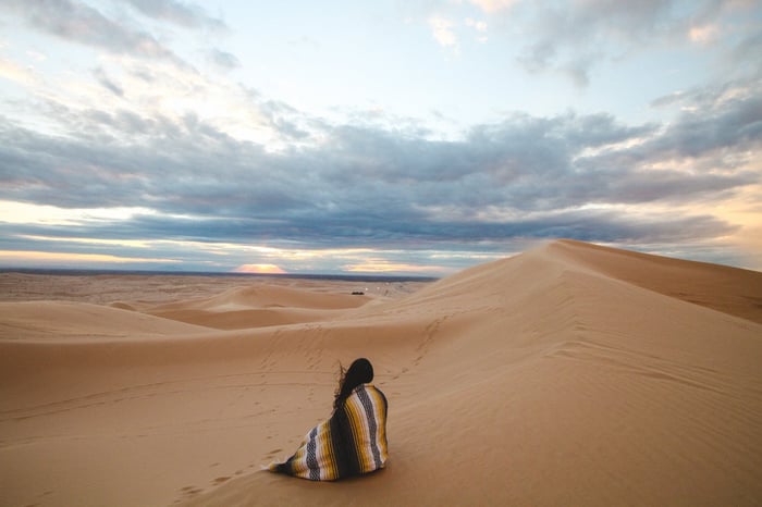 Woman wrapped in striped blanket sitting on desert sand dunes at sunset, symbolizing skin protection in harsh environments