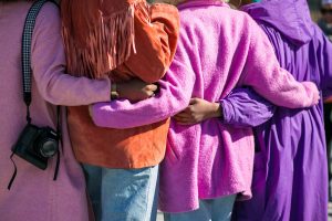 Three diverse women embracing in a supportive group hug wearing colorful sweaters