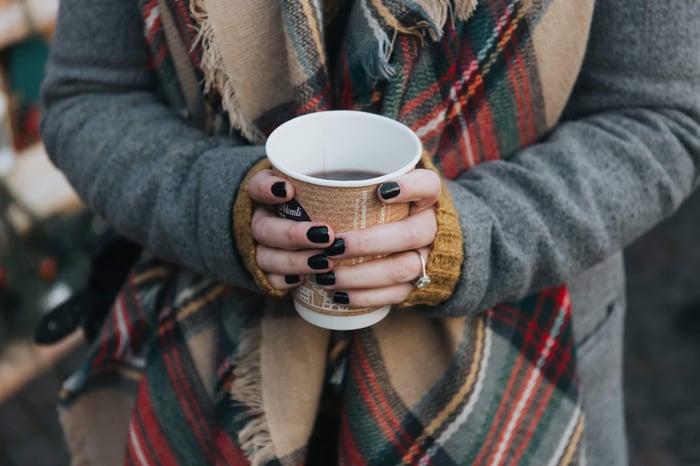 Woman in cozy grey sweater and tartan scarf holding a white mug with dark nail polish, perfect for demonstrating winter hand care