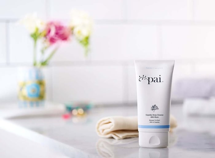 Pai Copaiba Deep Cleanse AHA Mask tube on marble surface with soft pink flowers in background