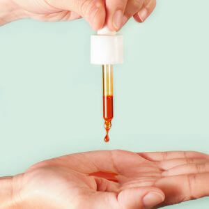 Hand squeezing dropper bottle releasing golden rosehip oil drop into cupped palm below