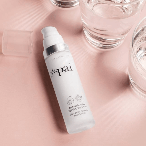 Pai Skincare Avocado & Jojoba Hydrating Day Cream bottle on soft pink surface with glass water vessels