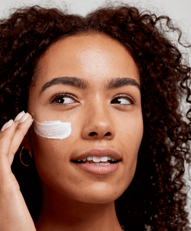 Woman with curly hair applying white facial cleanser to her cheek with fingertips, demonstrating gentle skincare routine