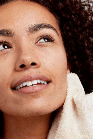 Woman with curly hair gently cleansing her face with a soft cream towel, demonstrating proper skincare routine