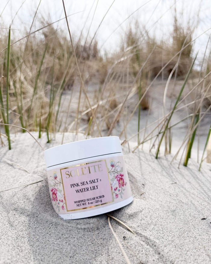 jar of pink sea salt body scrub nestled in the sand at the beach