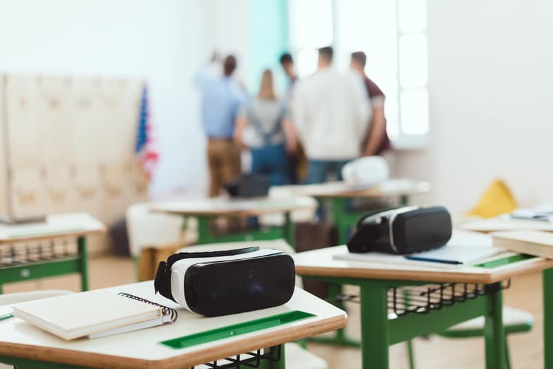 VR headsets and notebooks on school desks in a classroom, with students gathered near the windows.
