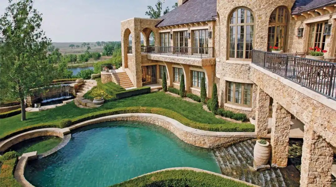 A grand, stone-clad mansion with a Mediterranean-inspired design sits atop a hill overlooking a serene landscape. The property features a cascading pool, lush gardens, and multiple terraces.