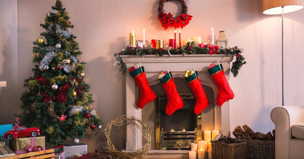 Christmas decoration ideas featuring a fireplace with candles and pine garland on the mantle, red socks hanging, candles on the floor next to baskets of chopped wood and pinecones, a decorated Christmas tree, and a wagon filled with gifts and toys