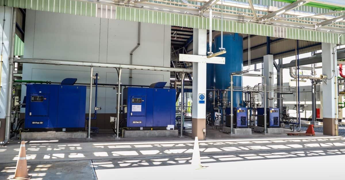 An industrial site with blue air compressor machines, a large blue tank, extensive piping, and a metal roof structure.