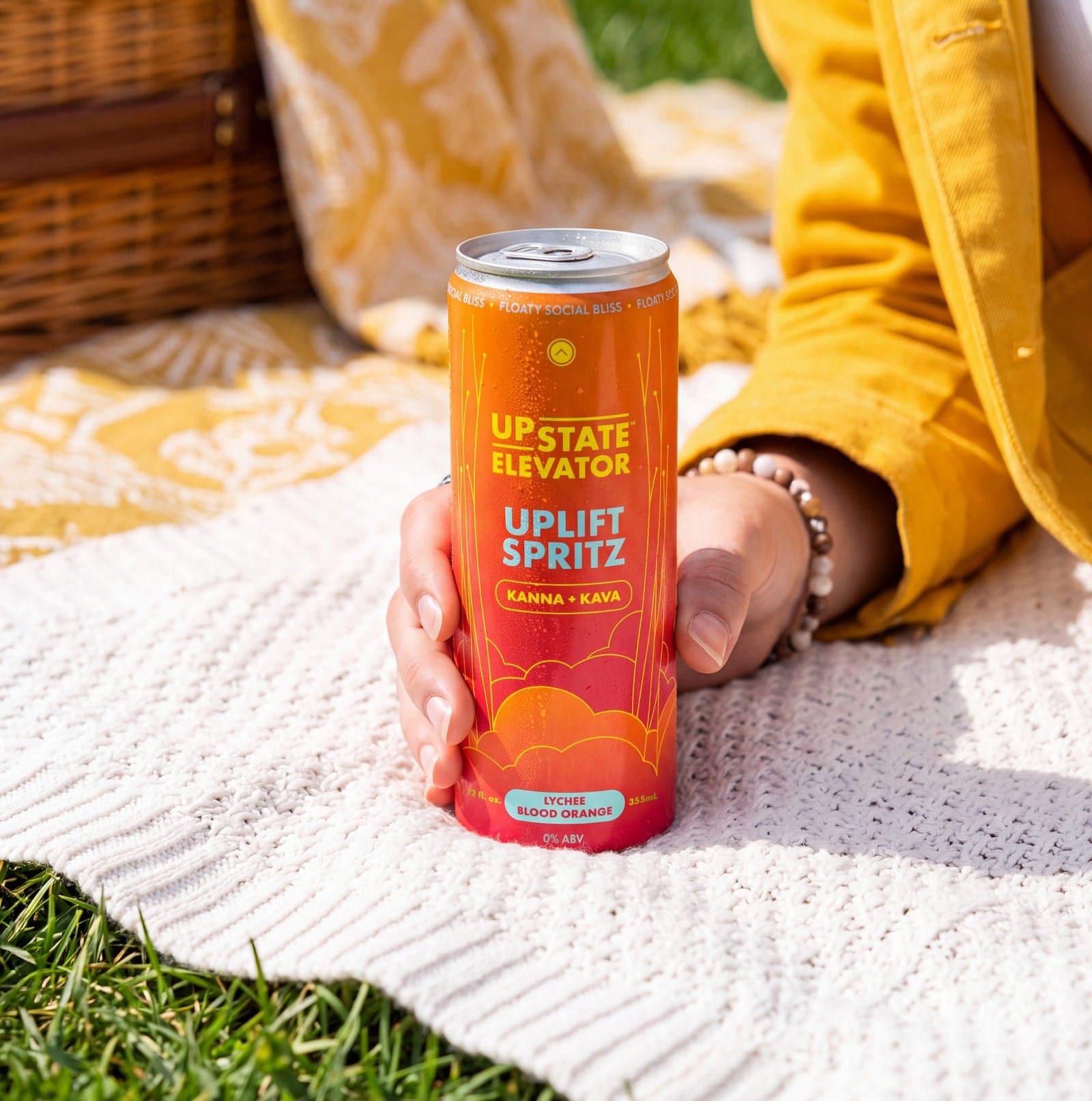 A hand holding an Upstate Elevator Lychee Blood Orange Uplift Spritz can on a white picnic blanket in the grass.