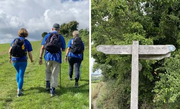 Three people hiking across green countryside with wooden walking signpost for South Downs Way