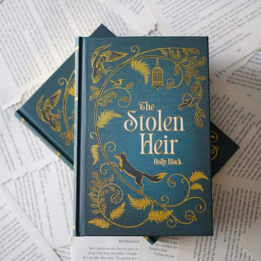 This image shows a close-up view of two hardcover books stacked on top of each other, placed on a surface covered with open pages of other books. The top book has a teal cover with an intricate golden design featuring leaves, birds, and a fox. The title "The Stolen Heir" is prominently displayed in large, golden letters, with the author's name "Holly Black" beneath it. The bottom book also has a teal cover with similar golden designs and the same title and author's name. The background consists of various pages of text, some of which are visible and partially legible.