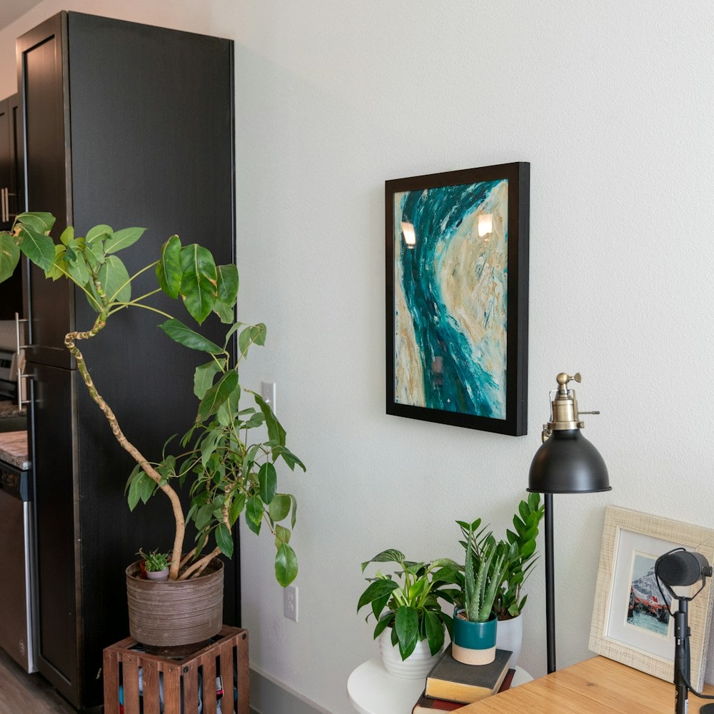 Black-framed teal artwork on a white wall beside houseplants and a desk lamp