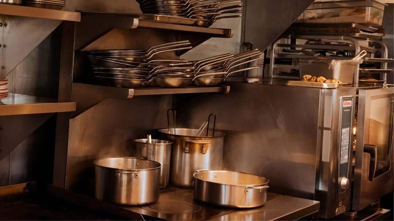 A commercial kitchen workspace features multiple stainless steel pots, pans stacked on shelves, and an oven, all arranged on a metallic surface.