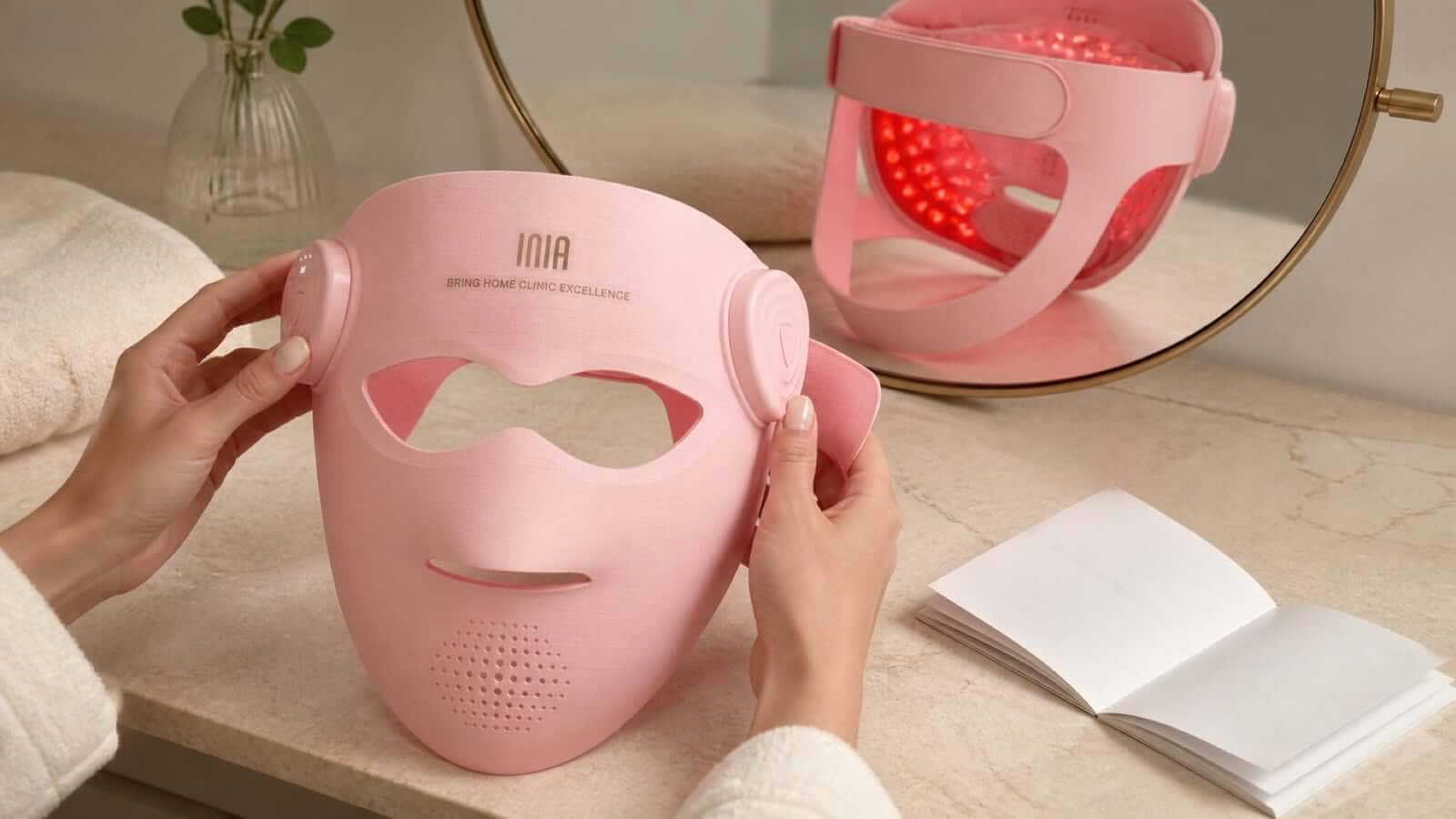 Hands inspecting a pink INIA LED face mask and its controls beside the instructions