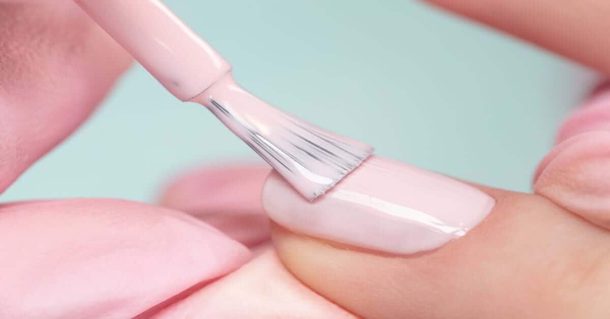 A pink nail brush applies soft pink polish to a fingernail, demonstrating professional technique.