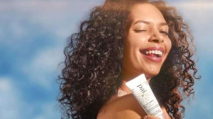 Woman with curly hair smiling joyfully while holding Pai SPF 30 sensitive sunscreen against blue sky background