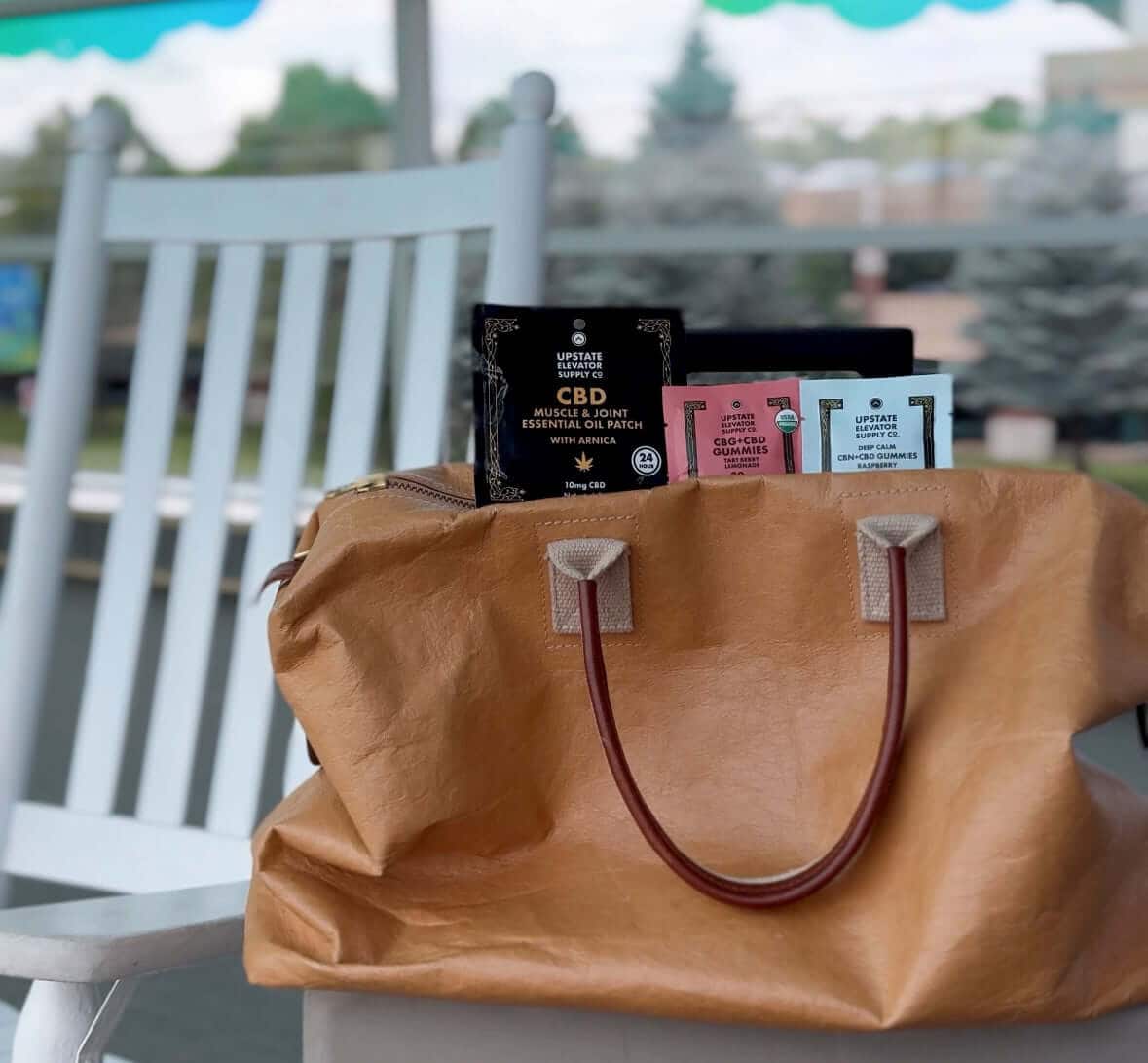 Tan tote bag holding an Upstate Elevator CBD muscle patch and gummy sample packs on a porch chair