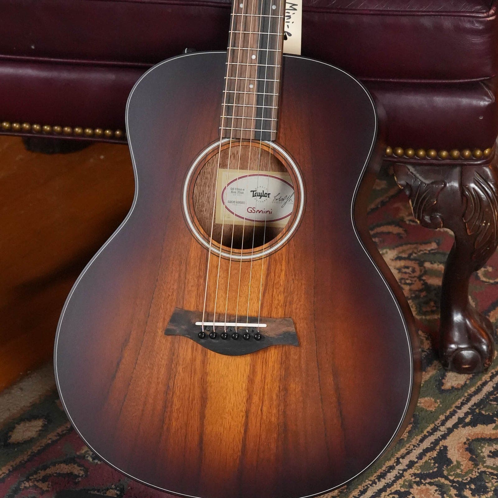 Taylor GS Mini-e Koa Plus in a shaded edgeburst finish on a rug in front of a leather chair