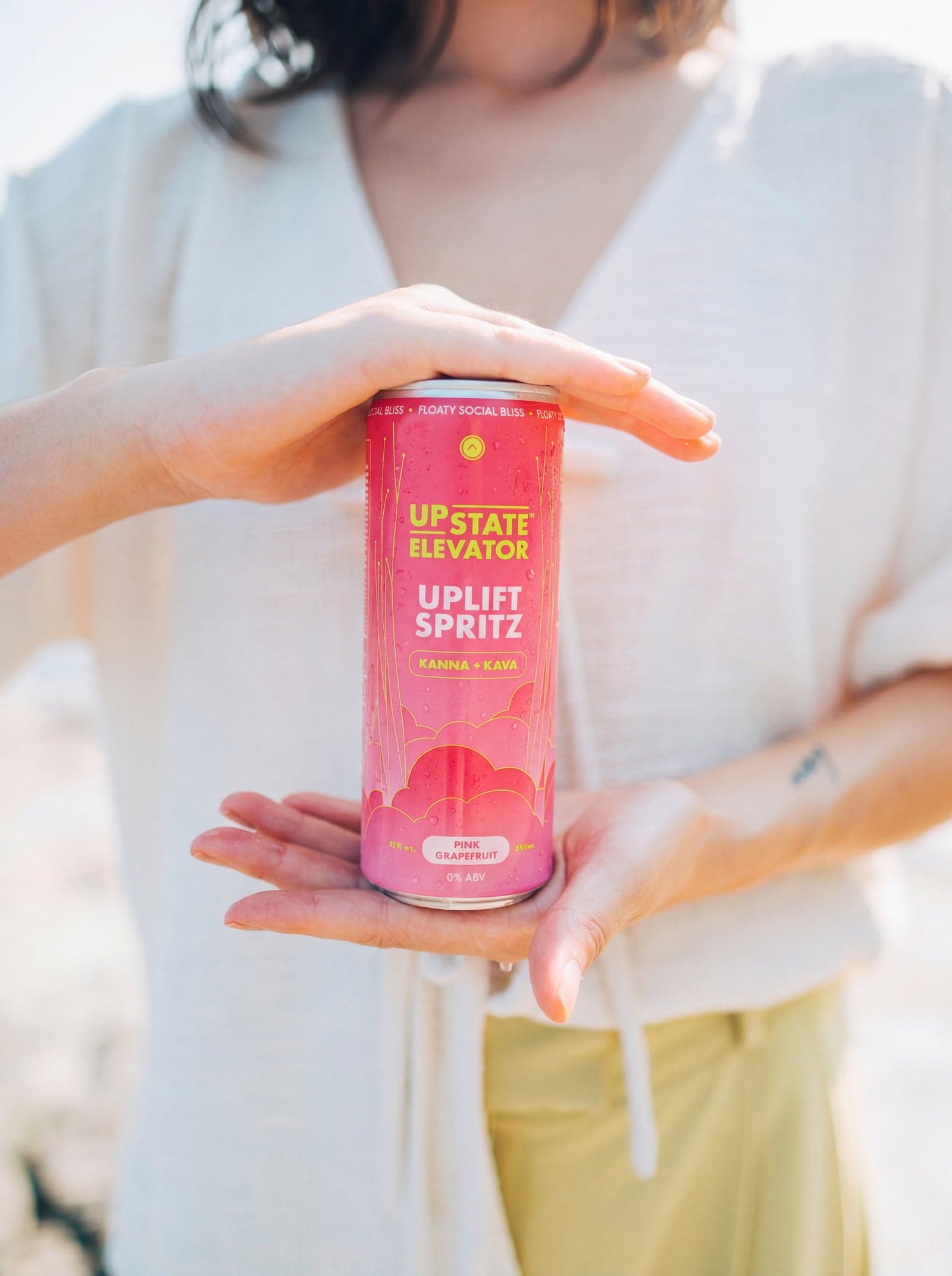 A woman holding an Upstate Elevator Pink Grapefruit Uplift Spritz can between her hands outdoors in bright sunlight.