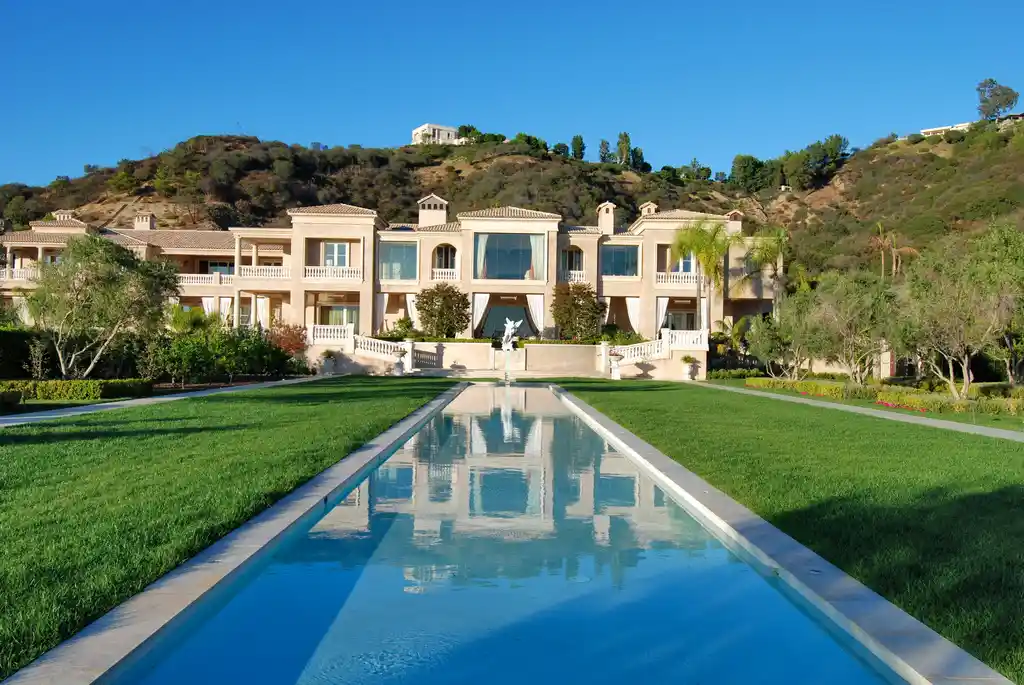 A grand, Mediterranean-style mansion sits atop a hill overlooking a lush, green landscape. The mansion features multiple levels, a large central courtyard, and a long, reflecting pool leading up to the entrance.
