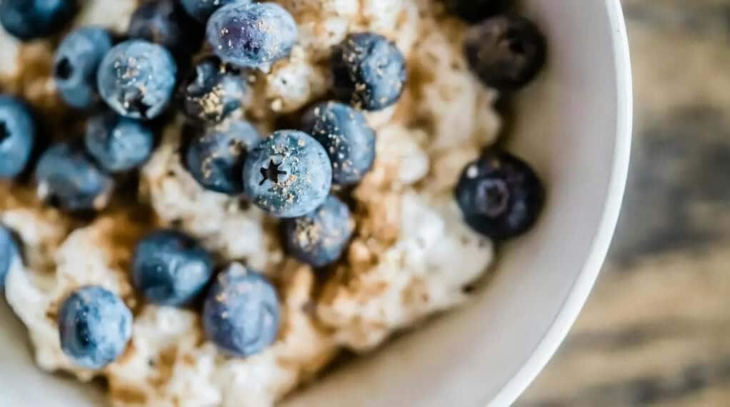 High protein meal prep of creamy blueberry oatmeal topped with fresh blueberries and flaxseed in a white bowl.