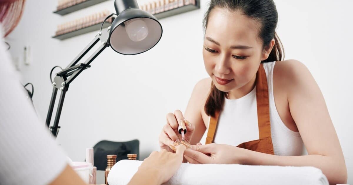A nail technician who is wearing an apron leans forward to apply polish on the nail of a sitting client in a salon.