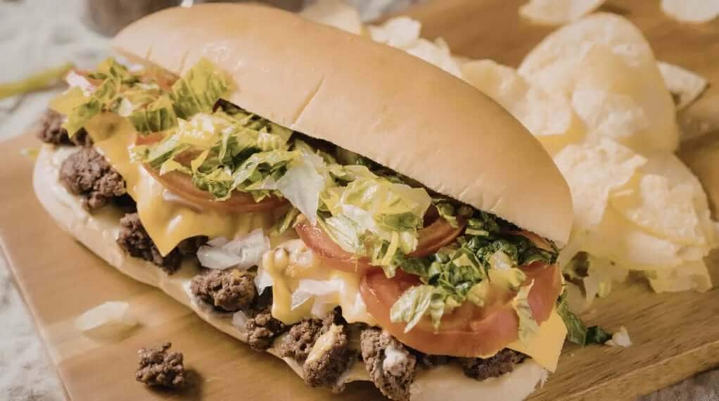 High protein meal featuring a loaded cheesesteak sub with seasoned ground beef, melted cheese sauce, sliced tomatoes, shredded lettuce, and diced onions on a soft hoagie roll.