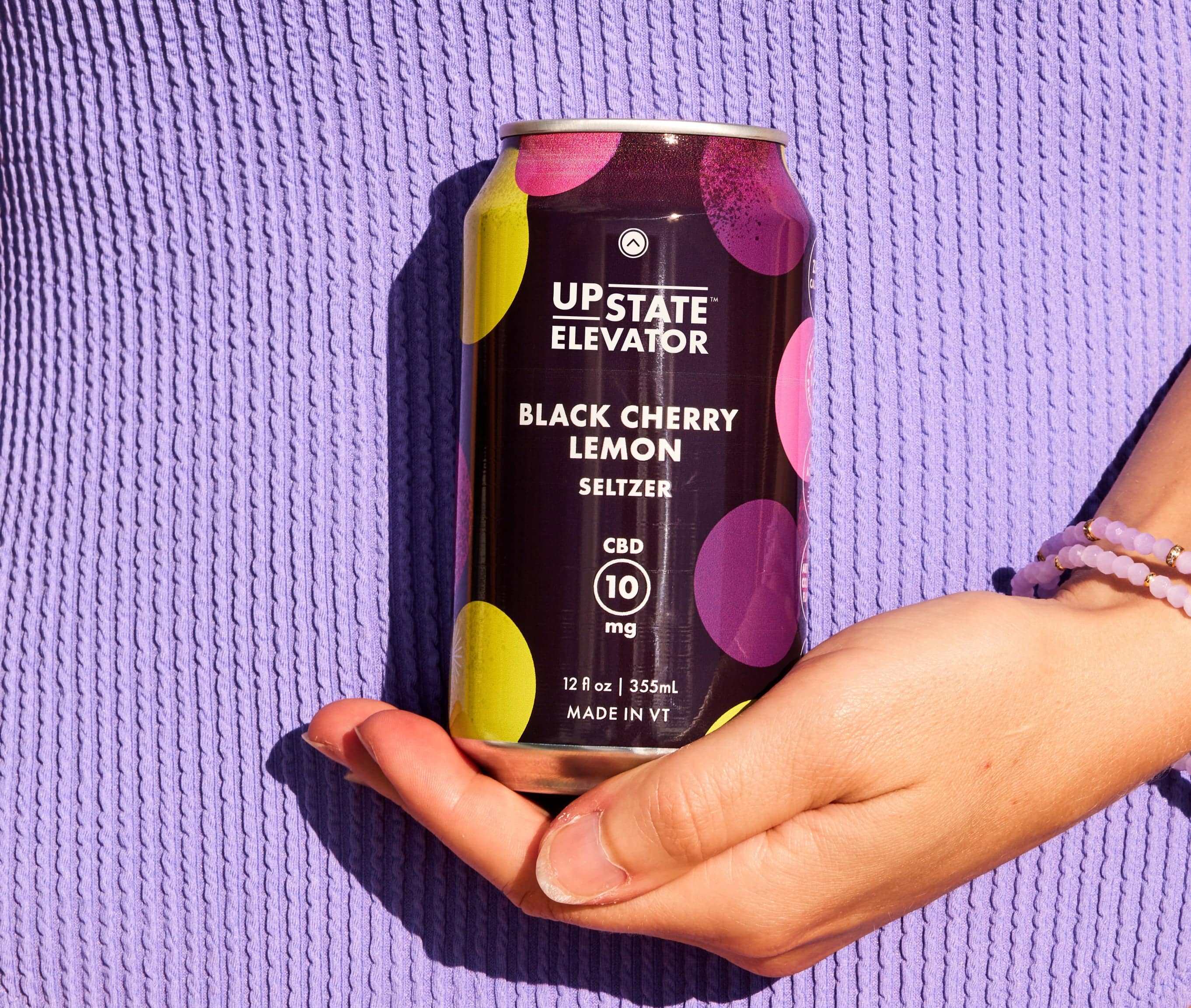 Hand holding an Upstate Elevator Black Cherry Lemon CBD seltzer can against a lavender textured background