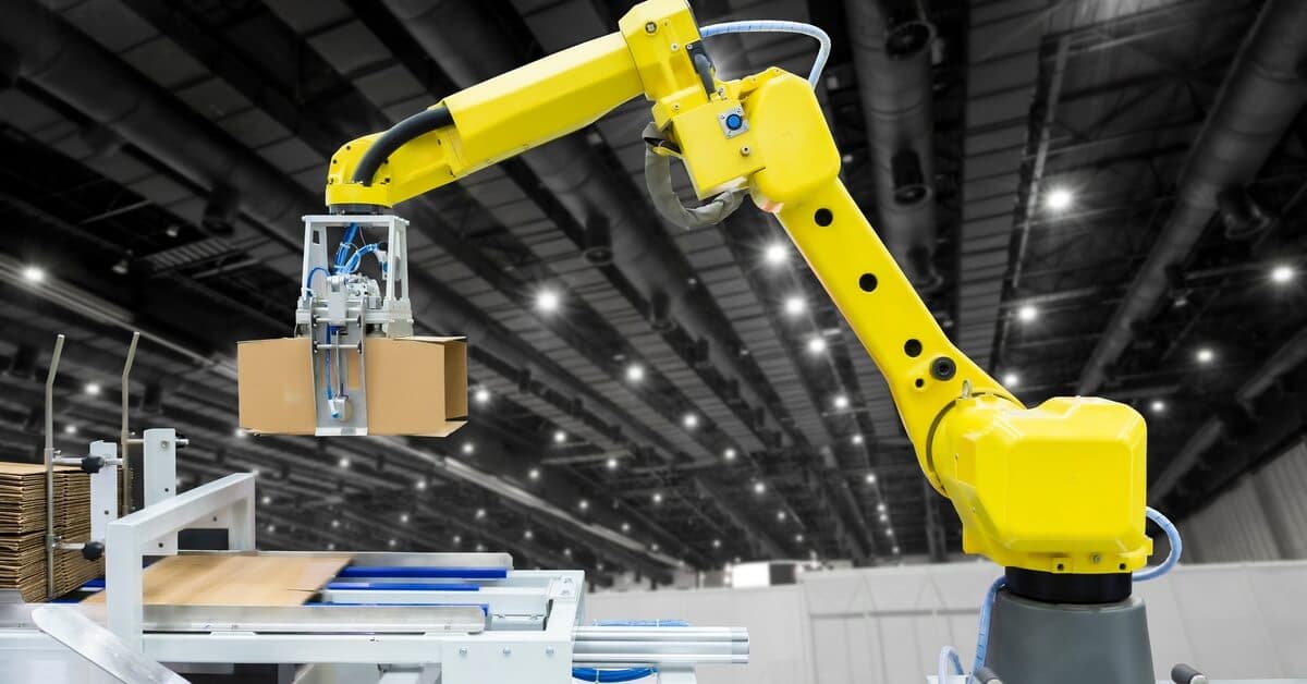 A yellow industrial robotic arm holds an assembled cardboard box in the air at a large warehouse facility.