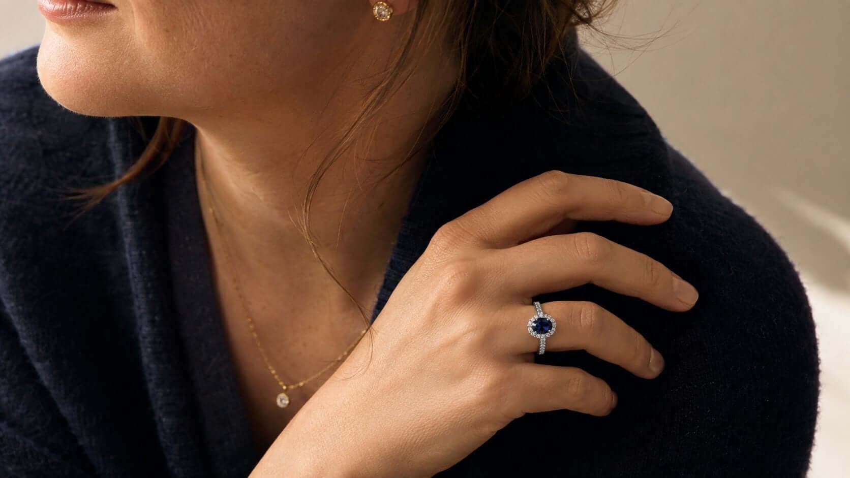 A blue sapphire and diamond ring worn with simple yellow-gold diamond jewellery and a restrained dark outfit.