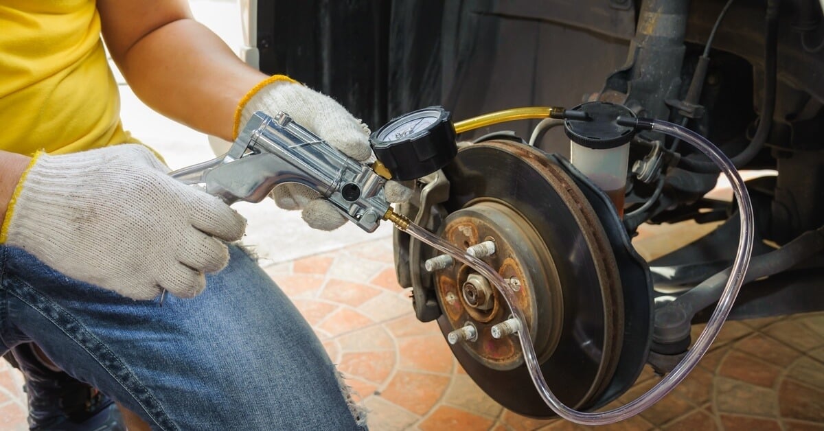 A person in a yellow shirt, jeans, and white gloves bleeding air from a vehicle's brake system using a specialized tool.