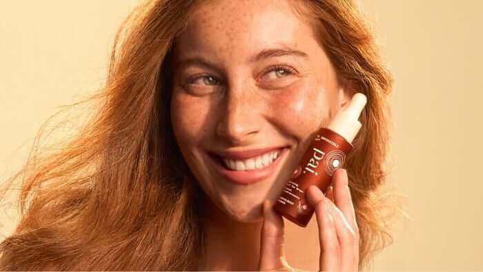 Smiling woman with freckled skin applying natural self tan drops to her face in warm golden lighting