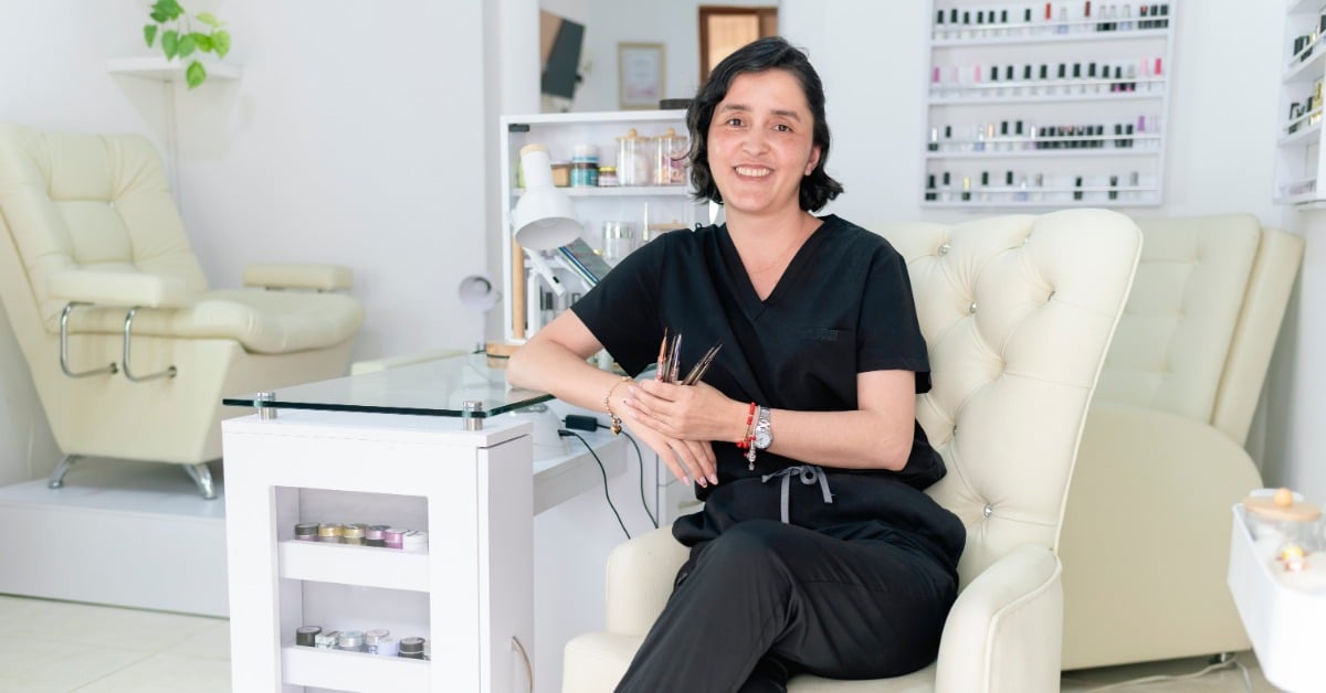 A smiling woman sits in a plush cream-colored chair and rests her elbow on a white desk with nail technician tools.