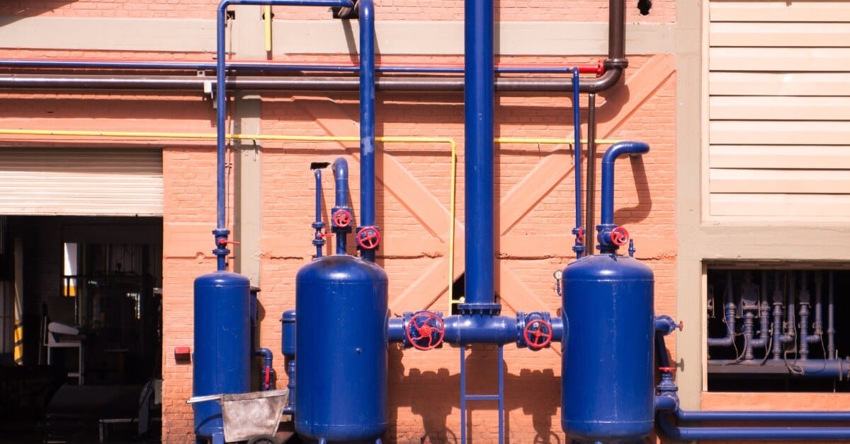 Three blue, vertical air compressor tanks, featuring several attached pipes, sitting outside a pink brick building.