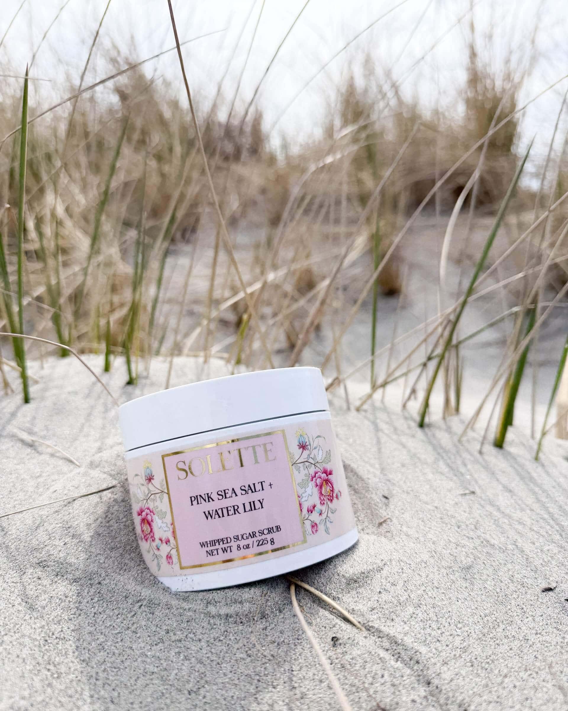 jar of pink sea salt body scrub nestled in the sand at the beach