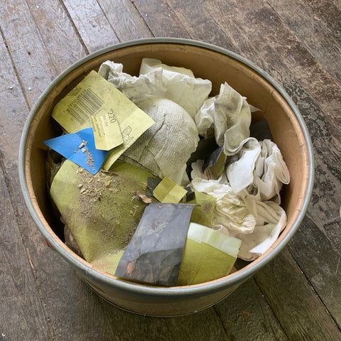 A refinery bucket filled with paper towels used sandpaper, and floor sweeps from a jewelry studio. The bucket is on a hardwood floor.