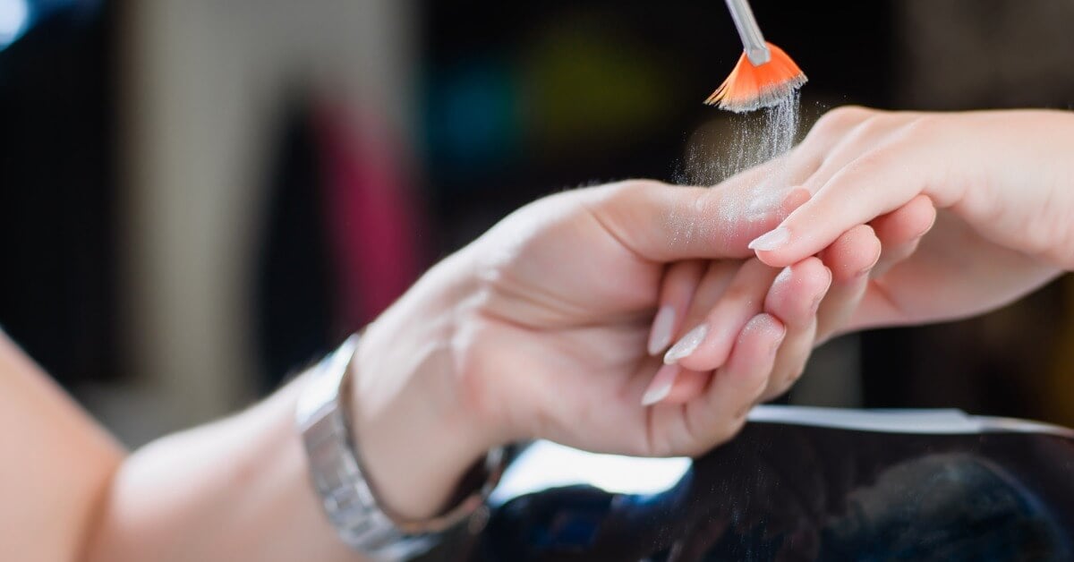 A nail technician holding the hand of a client and using a brush to drop silver nail powder over the nail surface.