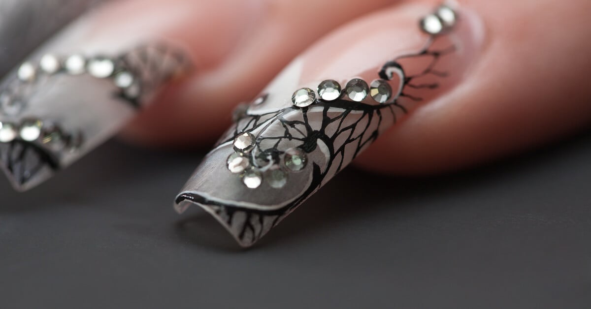 A close-up of a long acrylic nail on someone's hand. The nail features black fine-line designs and silver rhinestones.