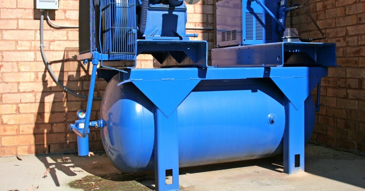 A large blue industrial air compressor with a horizontal receiver tank mounted outdoors against a brick wall.