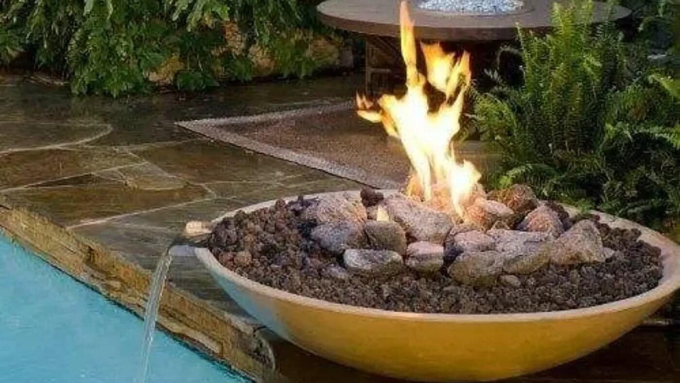 A close-up shot shows a round, light-colored fire bowl situated beside a pool. Flames rise from the center of the bowl, which is filled with dark lava rock and larger stones. A small spout on the side of the bowl releases a stream of water that flows into the pool. Lush green foliage surrounds the scene.