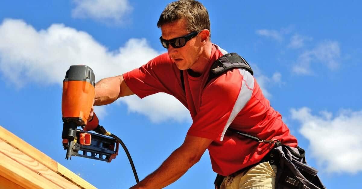 A man wearing a red shirt, khaki pants, and sunglasses using an orange nail gun to nail pieces of wood outside.