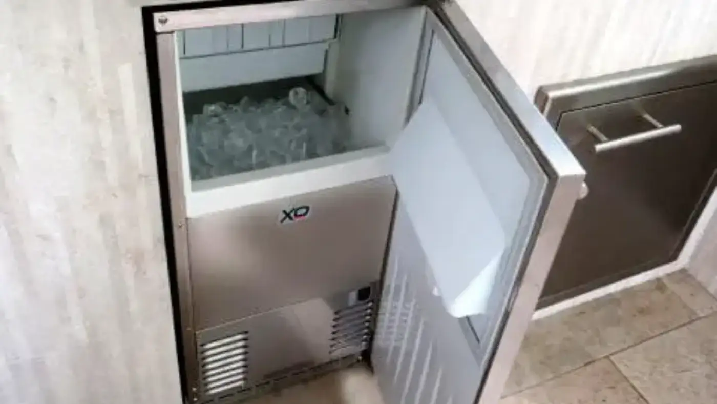 Stainless steel under-the-counter ice maker built into an outdoor kitchen. The door is open, revealing a bin full of ice cubes.